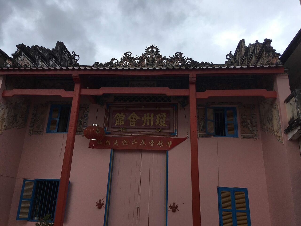 The pink front of a Chinese temple with a decorated tiled roofline, red pillars and a sign in Chinese characters