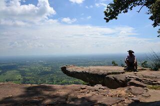 A lone figure sits on a rock ledge overlooking a wide green plain under a cloudy sky.