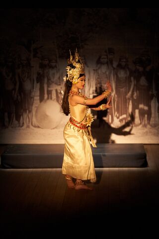 A Khmer classical dancer in a golden crown and costume performing on a dark stage under a spotlight.
