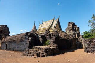 A golden-roofed modern Buddhist hall standing among ruined laterite walls on sandy ground.