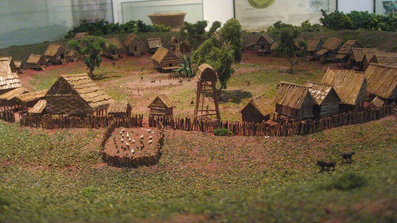 A model of thatched houses inside a round enclosure of banks and a fence, on a green landscape.