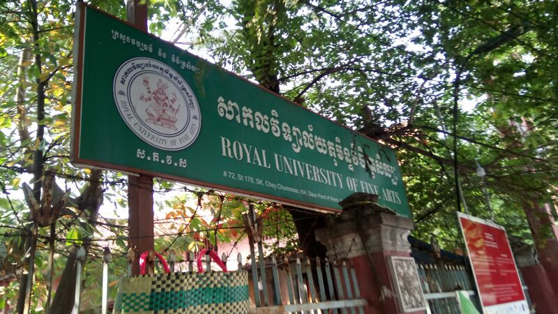 A green signboard with the university's seal and its name in Khmer and English above a gate under trees.
