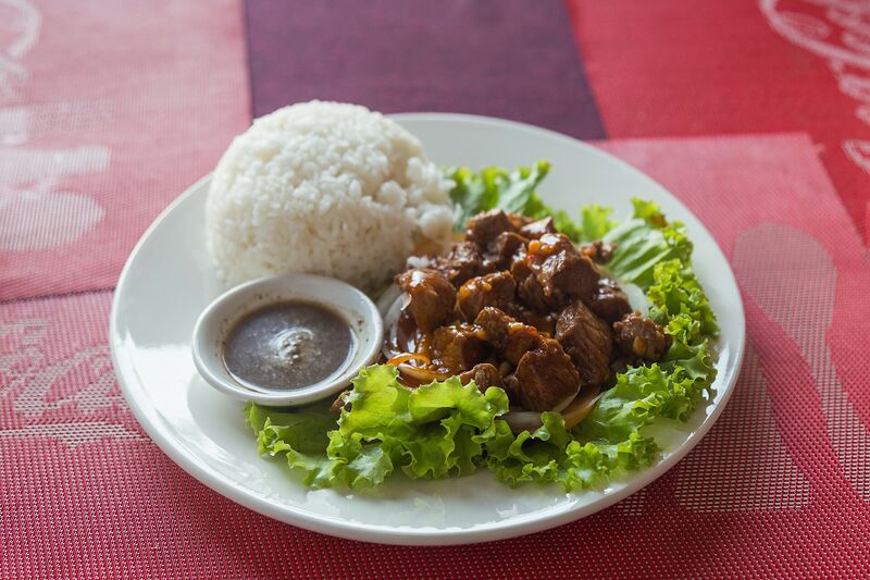 Cubes of glazed beef on lettuce beside a mound of white rice and a small bowl of dark pepper sauce.