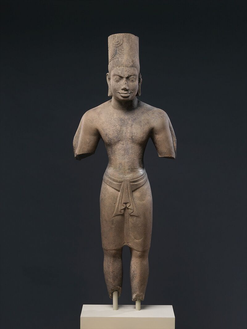 A standing stone figure with a tall headdress, its arms broken off, on a plain base against a dark background.