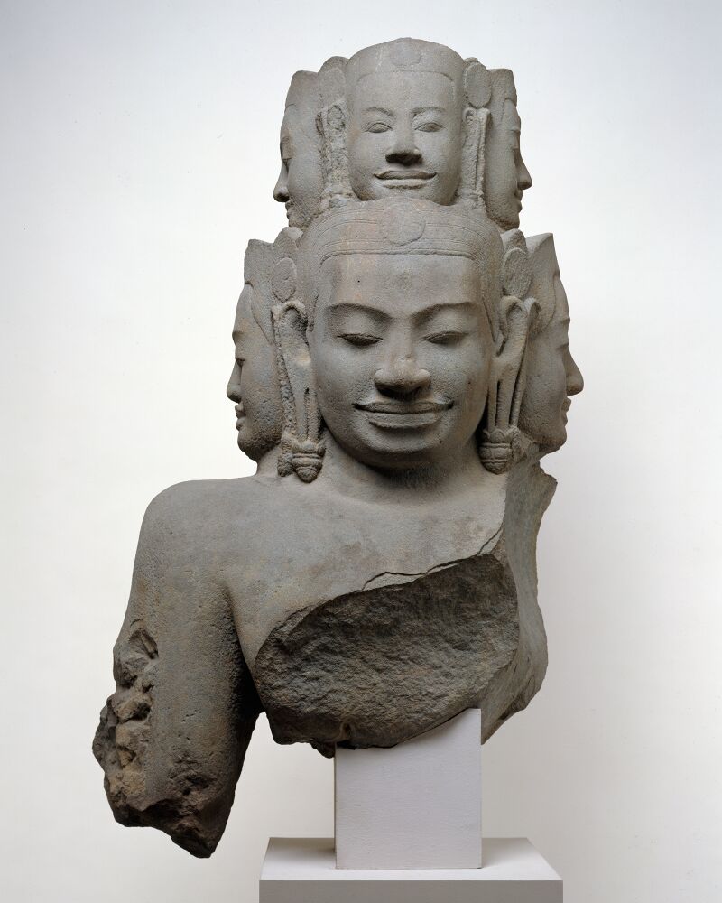 A gray sandstone bust with many crowned heads in tiers, smiling serenely, mounted on a white base.