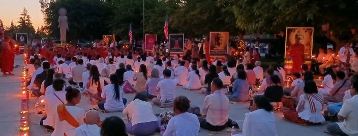 Rows of people in white seated on mats at dusk holding candles, monks and Buddhist banners at the front, a tall standing Buddha statue behind.