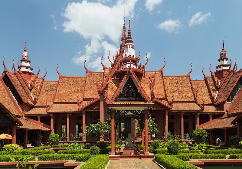 A red building with layered, steeply pitched roofs and tall spires, behind a garden of clipped hedges and a central statue.