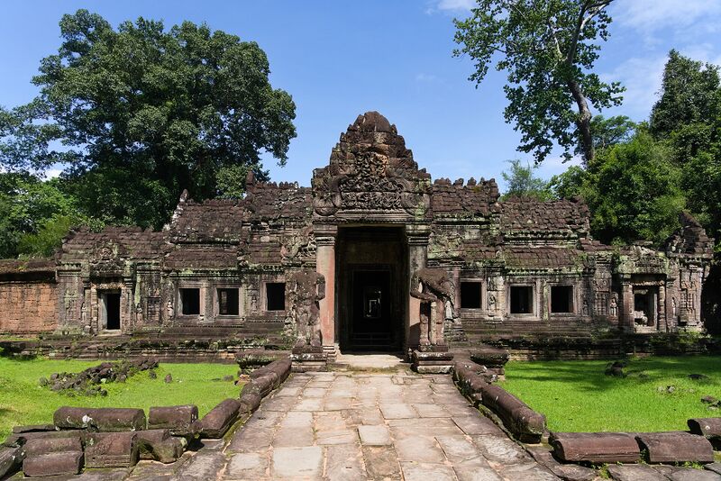 A long, weathered stone gallery with a carved central doorway, reached by a stone path between lawns and tall trees.