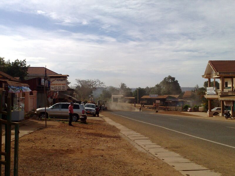 A wide road with a dusty haze ahead, low shops and parked cars on the left, and a tiled-roof building on the right.