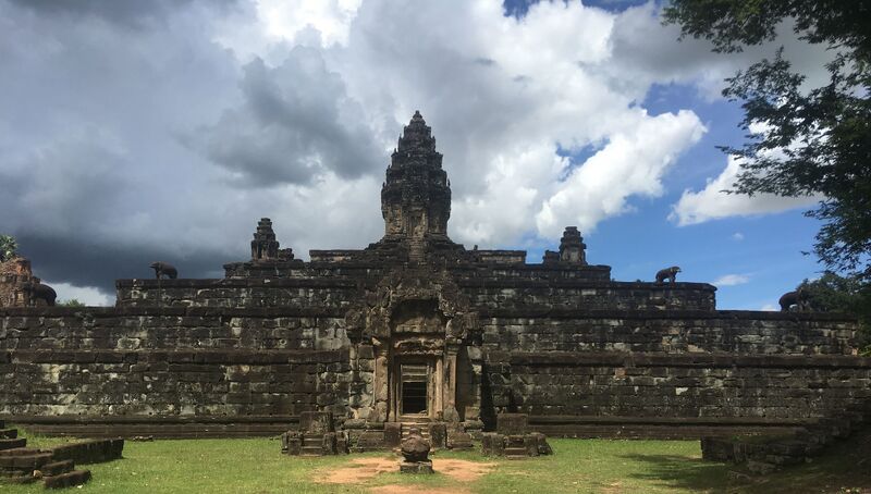 A stepped pyramid of dark gray stone rises in broad terraces to a single tall tower, with smaller towers at its corners and stone elephants standing on the terrace edges. A grassy path with scattered stone blocks leads to a doorway in the base, under a sky of heavy gray and white clouds.