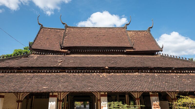 A Khmer temple hall with a long, low red-tiled roof in two tiers under a blue sky.