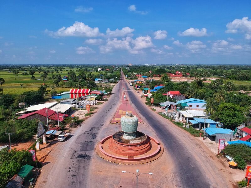 Seen from above, a large carved green-gray bowl stands on a round red base in the middle of a straight divided road. Shops with colored metal roofs and stone statues line both sides, with rice fields and palm trees beyond under a blue sky with white clouds.
