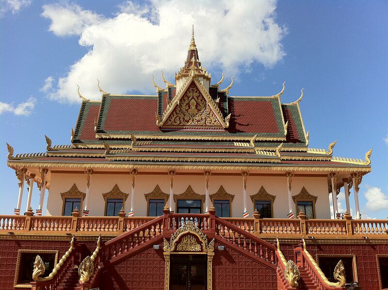 Khmer-style temple hall with tiered red roofs, gold trim and ornate front stairway under a blue sky.