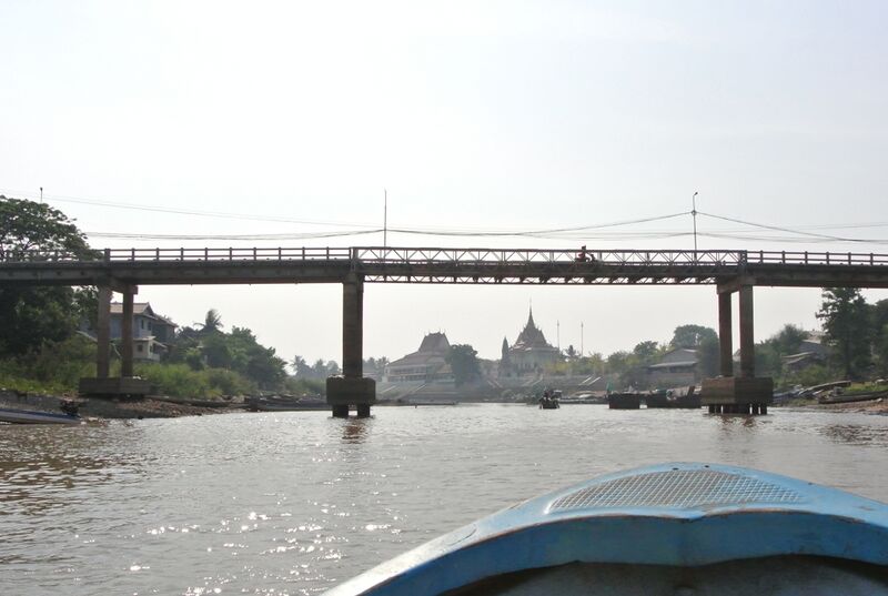 A wide canal runs toward a long concrete footbridge, with a pagoda roof and low buildings behind it and the bow of a blue boat in the foreground.