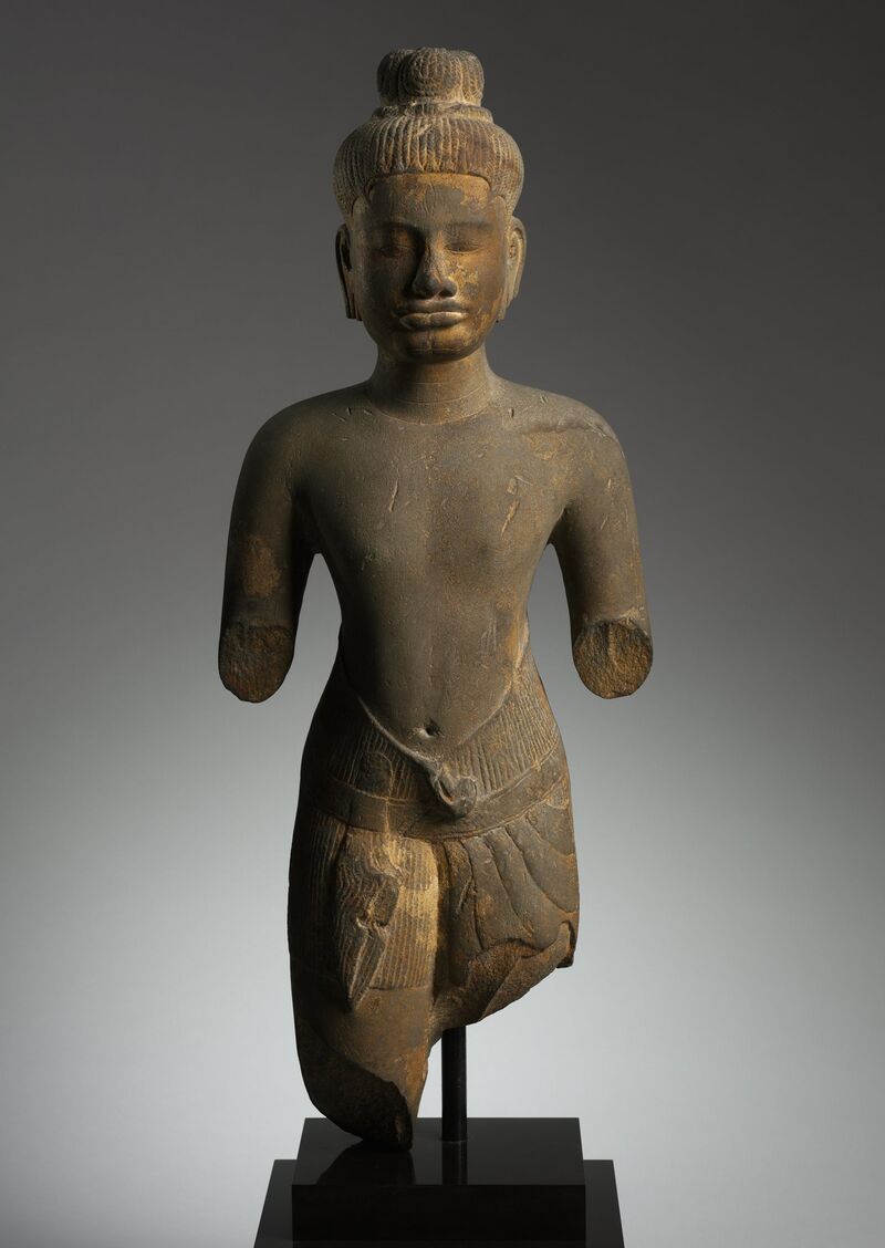A gray sandstone torso of a standing god with hair in a tall tied bun, a calm face with a pointed beard, and a short pleated garment with a low belt, its forearms and lower legs broken away.