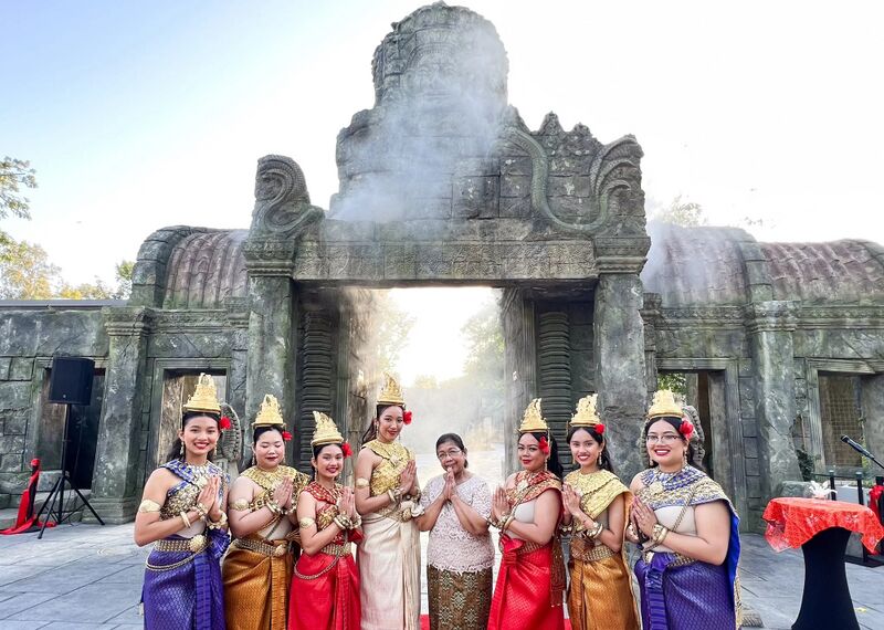 Eight dancers in Khmer classical costumes and an older woman stand with palms joined before a stone gate carved with a face tower.
