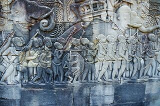 A gray sandstone relief of rows of marching soldiers with spears, shields and an elephant.