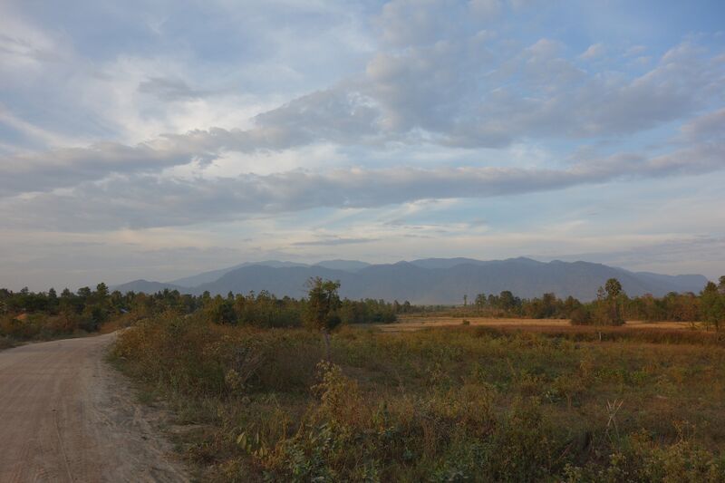 Layers of blue-gray forested ridges stretch across the horizon under a wide sky with long bands of cloud. In front, a dirt road curves past dry grass, scrub and scattered trees.