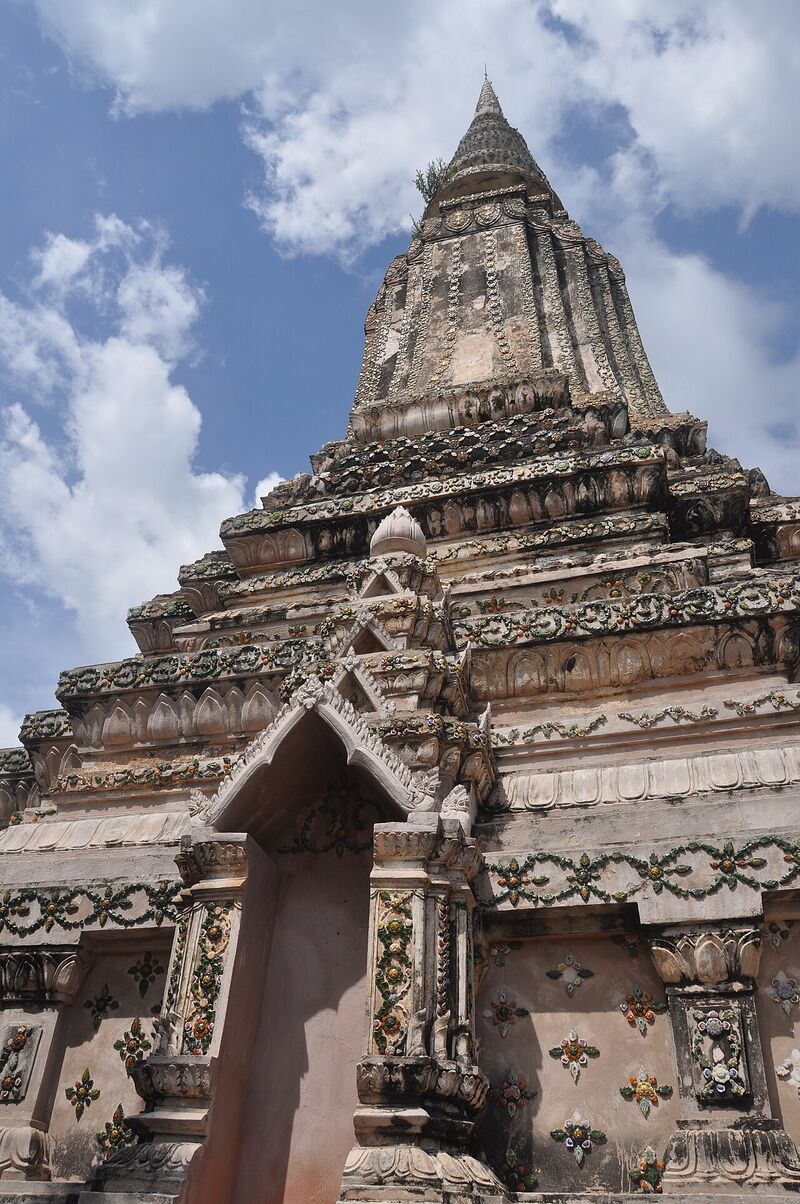 A tall weathered stone stupa with tiered carved bands and a pointed spire, under a blue sky with white clouds.