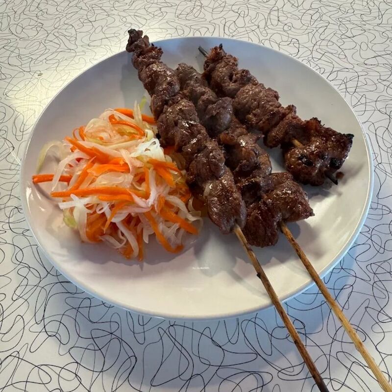 Two grilled beef skewers on a white plate next to pickled shredded carrot and papaya.