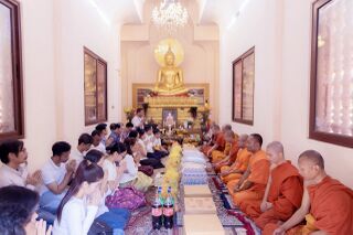 Monks in orange robes seated along one side of a long hall facing lay people, with a Buddha image on the altar at the end.
