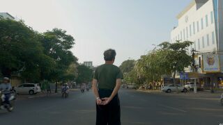 A man seen from behind stands with his hands clasped in the middle of a city street lined with trees and buildings.