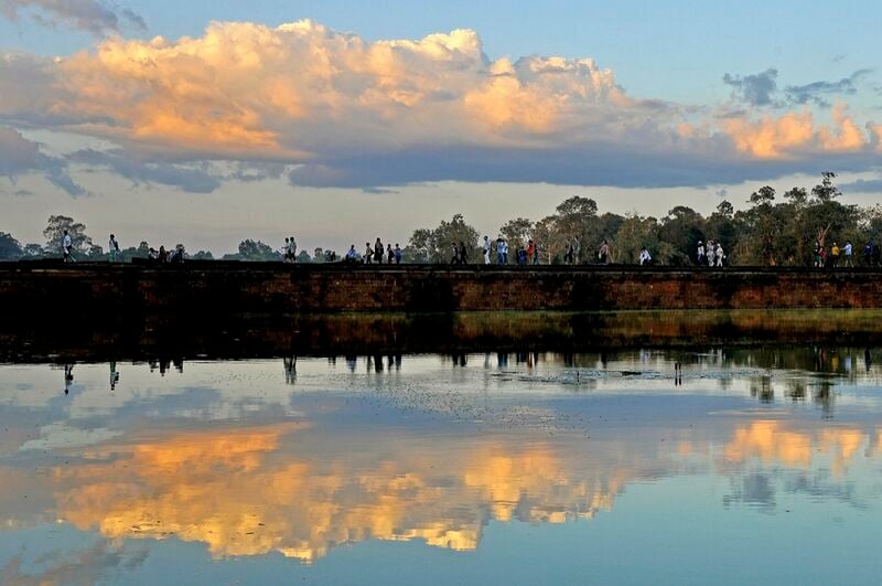 Orange and gold clouds over the long Angkor Wat causeway and moat at sunset, with visitors walking along the dark stone wall as silhouettes.