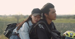 A young man rides a motorbike along a rural road with a young woman in a cap leaning against his back.