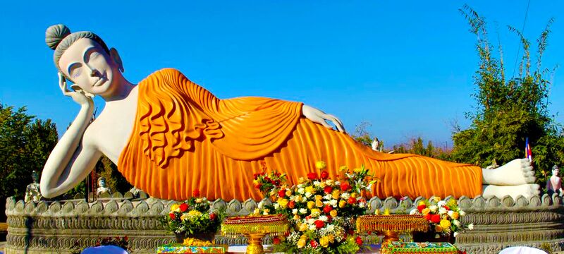 A large white reclining Buddha in an orange robe on a carved base, with flower offerings in front and a clear blue sky.