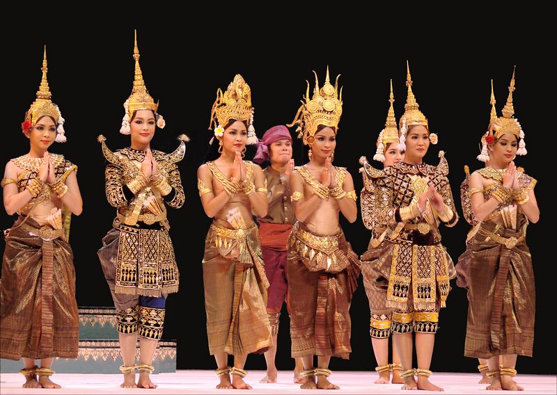 Dancers in gold crowns and costumes stand in a row on stage with palms pressed together.