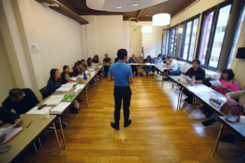 A teacher stands in a long classroom with wooden floors while adult students sit at tables along both sides.
