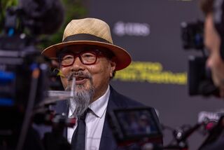 A bearded man in a straw hat and glasses smiling on a red carpet, with a camera beside him.