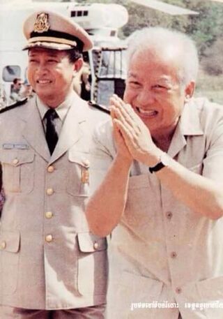An older man in a light shirt with palms pressed together at chest height, beside a smiling man in a beige uniform.
