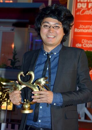 A man in glasses and a dark jacket holds a gold swan-shaped trophy outdoors.