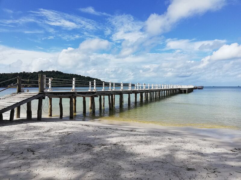 A long wooden pier runs out over shallow turquoise water from a white sand beach under a blue sky.