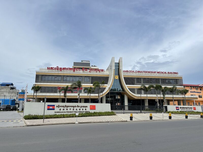 A modern hospital building with a curved glass entrance and red Khmer and English lettering along its roof, behind a low wall with Cambodian and Chinese flags.