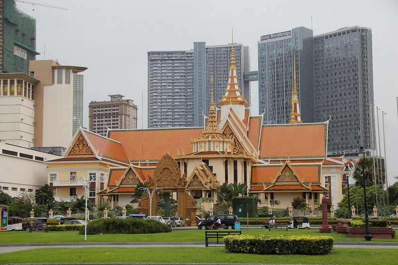 A low Khmer-style building with orange tiled roofs and a gilded multi-tiered spire at its center, set behind a green lawn and hedge with tall towers rising in the background.