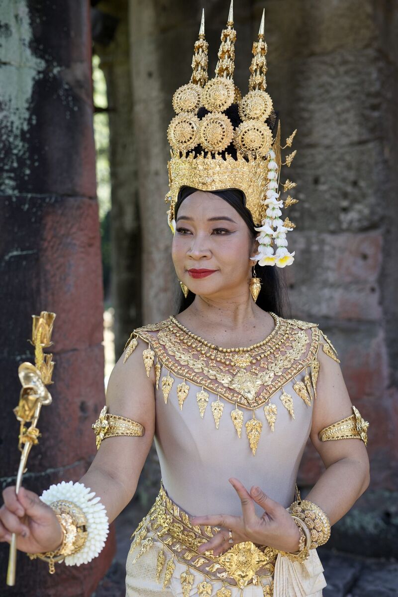 A dancer in a gold apsara costume and tall crown holds a golden flower, standing among stone temple pillars.