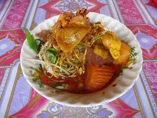 A white bowl of rice noodles in red curry topped with chicken, carrot, crisp vegetables and herbs, on a patterned cloth.
