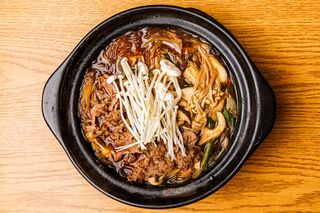 A black stone pot of beef and mushrooms in broth topped with enoki, seen from above.