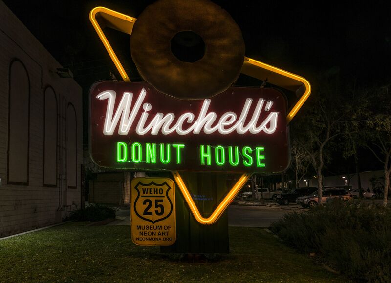 A lit neon sign shaped like a triangle with a large donut on top, at night.
