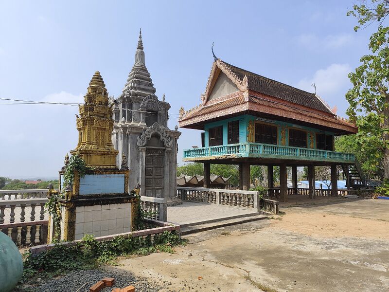 A gold shrine and a gray stupa beside a turquoise hall raised on stilts, under a blue sky.