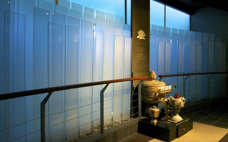 Backlit glass panels engraved with names behind a wooden rail, with a bronze urn and lotus offering vessel in front.