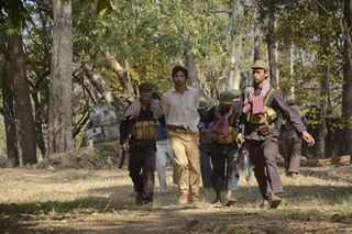 Armed soldiers in black escort a man in a white shirt through a dry forest.
