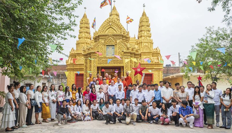 A large group of monks and lay people in white and traditional dress posing before a gilded Khmer-style temple with three towers.