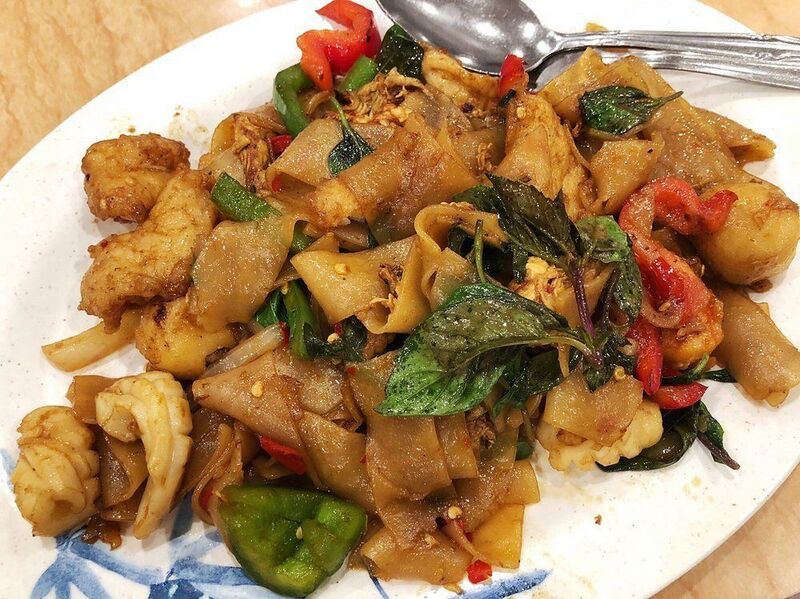 A white plate of wide stir-fried noodles with meat, peppers, tomato and basil leaves.