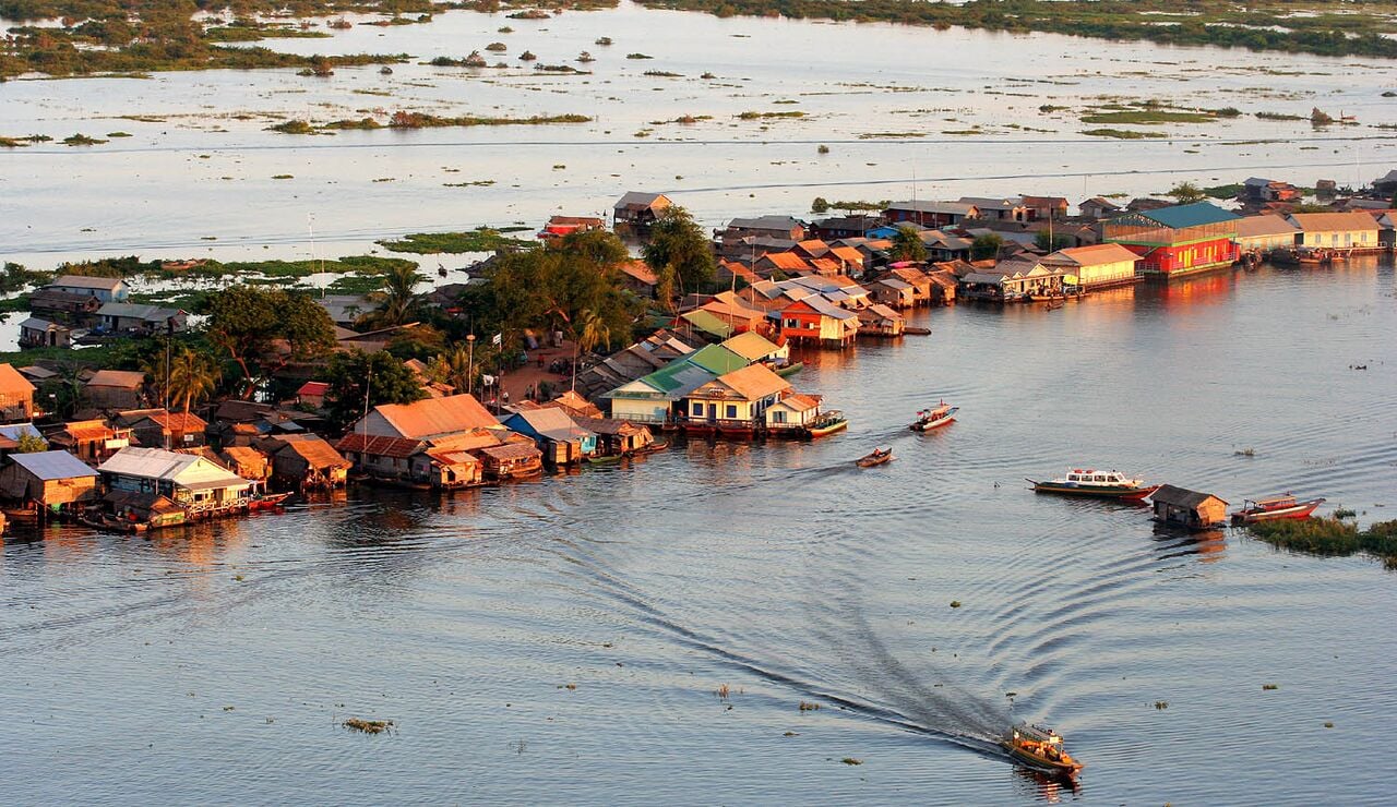 Rows of floating houses with orange and blue roofs seen from above on brown lake water, with a boat leaving a wake