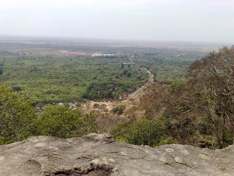 A wide flat green plain stretching to a hazy horizon, seen from a rocky hilltop.