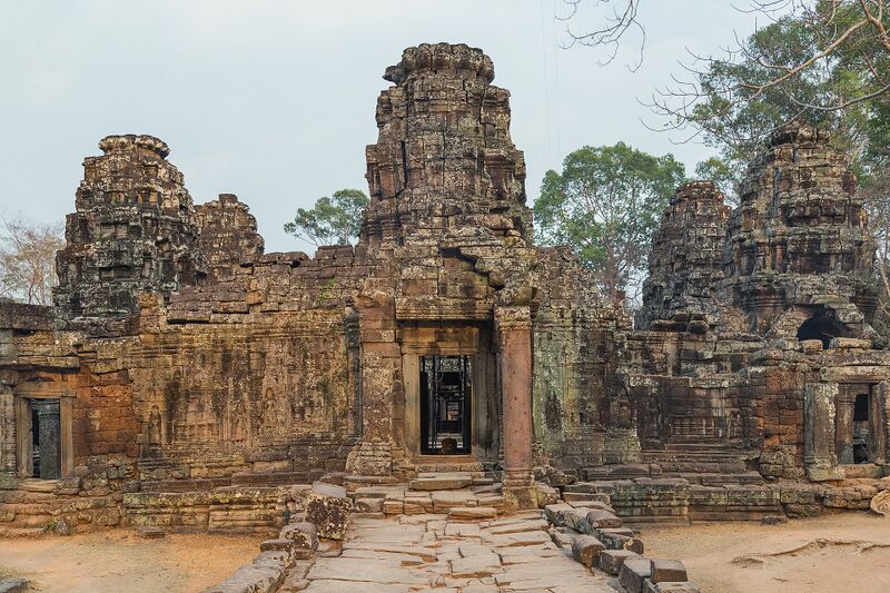 A ruined sandstone temple with several towers and a long gallery, set on bare ground with trees behind.