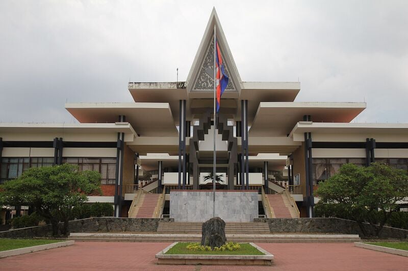A wide modern building with layered flat roofs under a tall pointed gable, a flag in front, and wide steps leading to its entrance